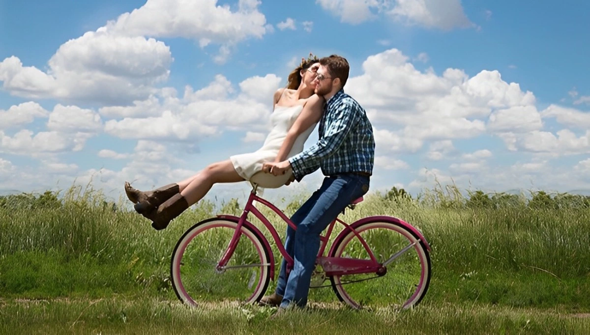 un homme a chevauche un velo rouge et une femme assise sur le guidon l embrasse sur la joue dans un champs sur fond de ciel bleu et des nuages blancs eparcemes