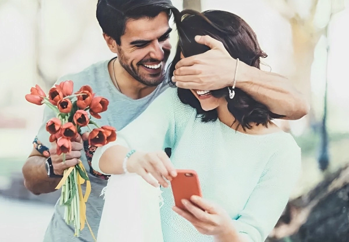 un homme souriant fait une surprise a une femme tenant un telephone avec un bouquet de fleurs rouges dans une main et avec l autre couvre ses yeux